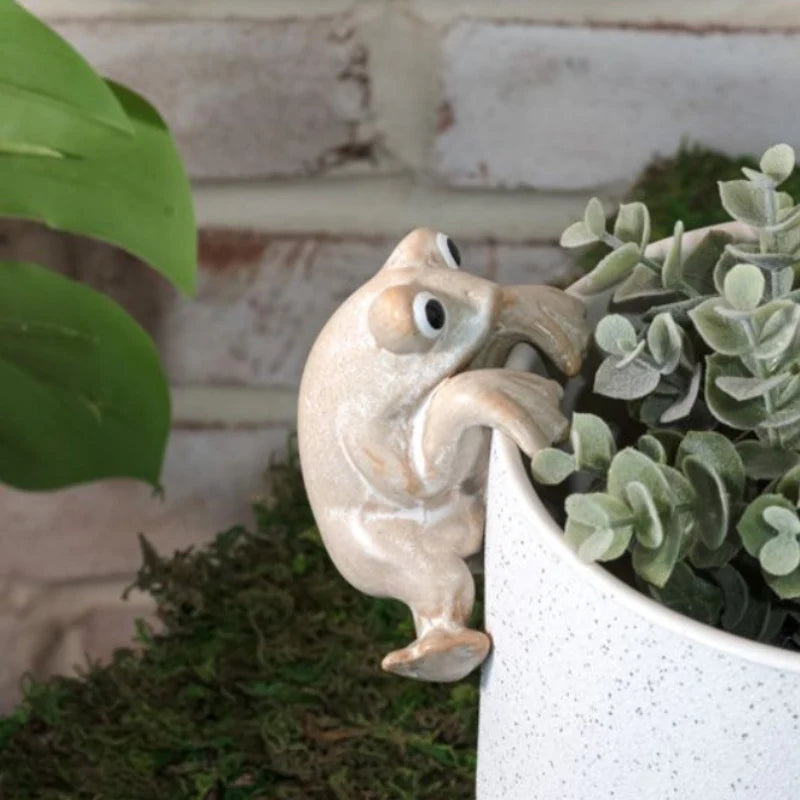 Beige ceramic frog figurine hanging on edge of a white planter