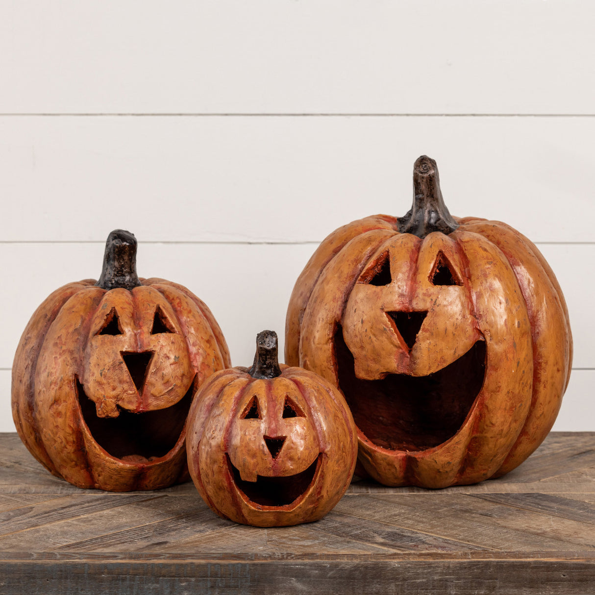 A set of three resin mache pumpkins in varying sizes with a smiling face design, placed on a wooden surface against a white background.