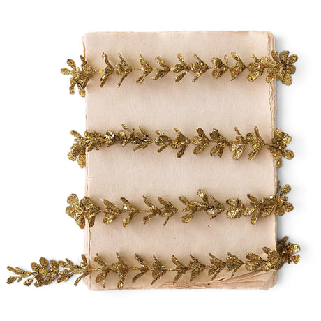 Slender gold glitter faux boxwood garland with small leaves, arranged in loose rows across an ivory background.