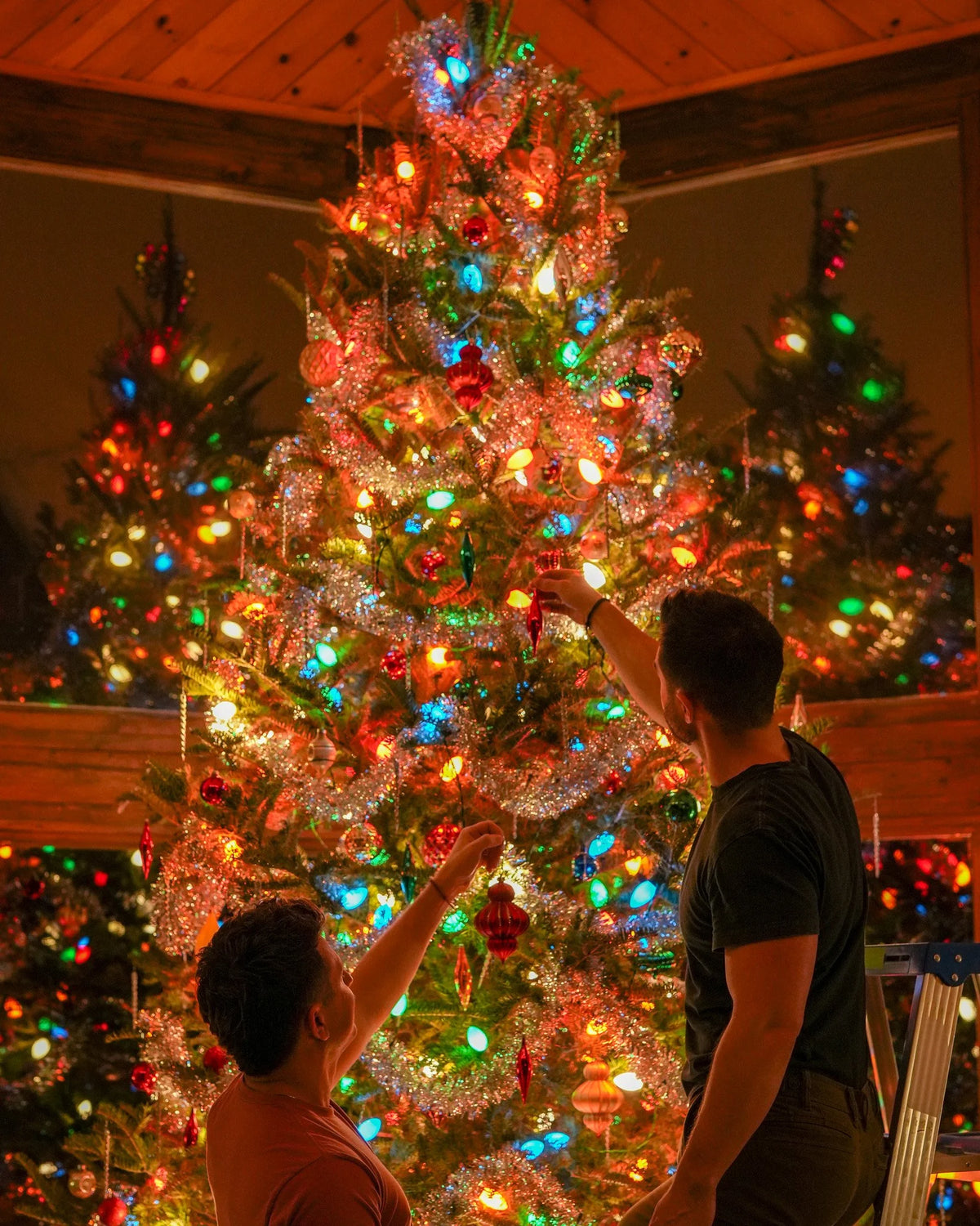 Two people decorating a Christmas tree with multicolor Tru-Tone lights, silver tinsel, and ornaments.